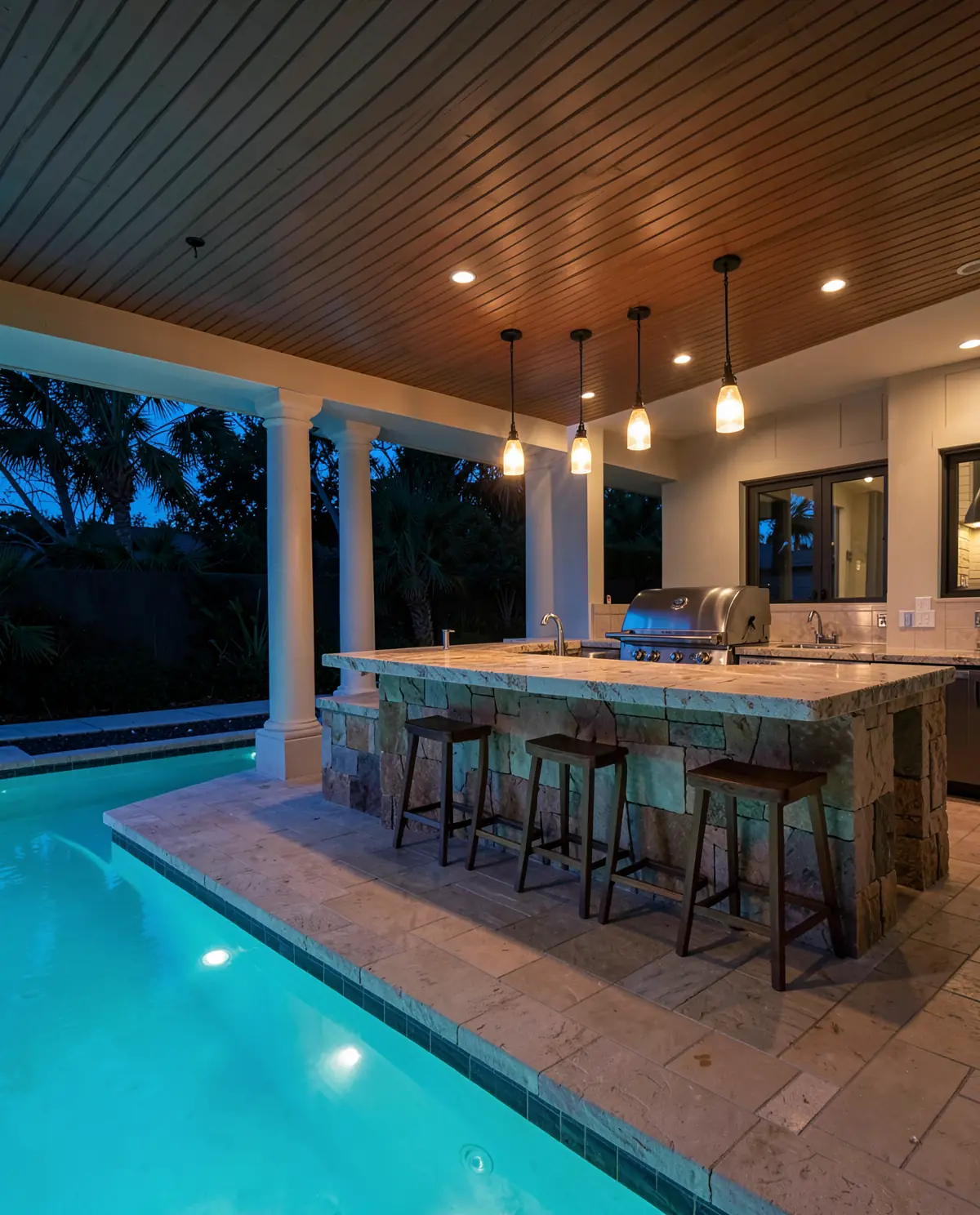 An outdoor kitchen under a lit lanai beside a pool at dusk (illustrative, generated).